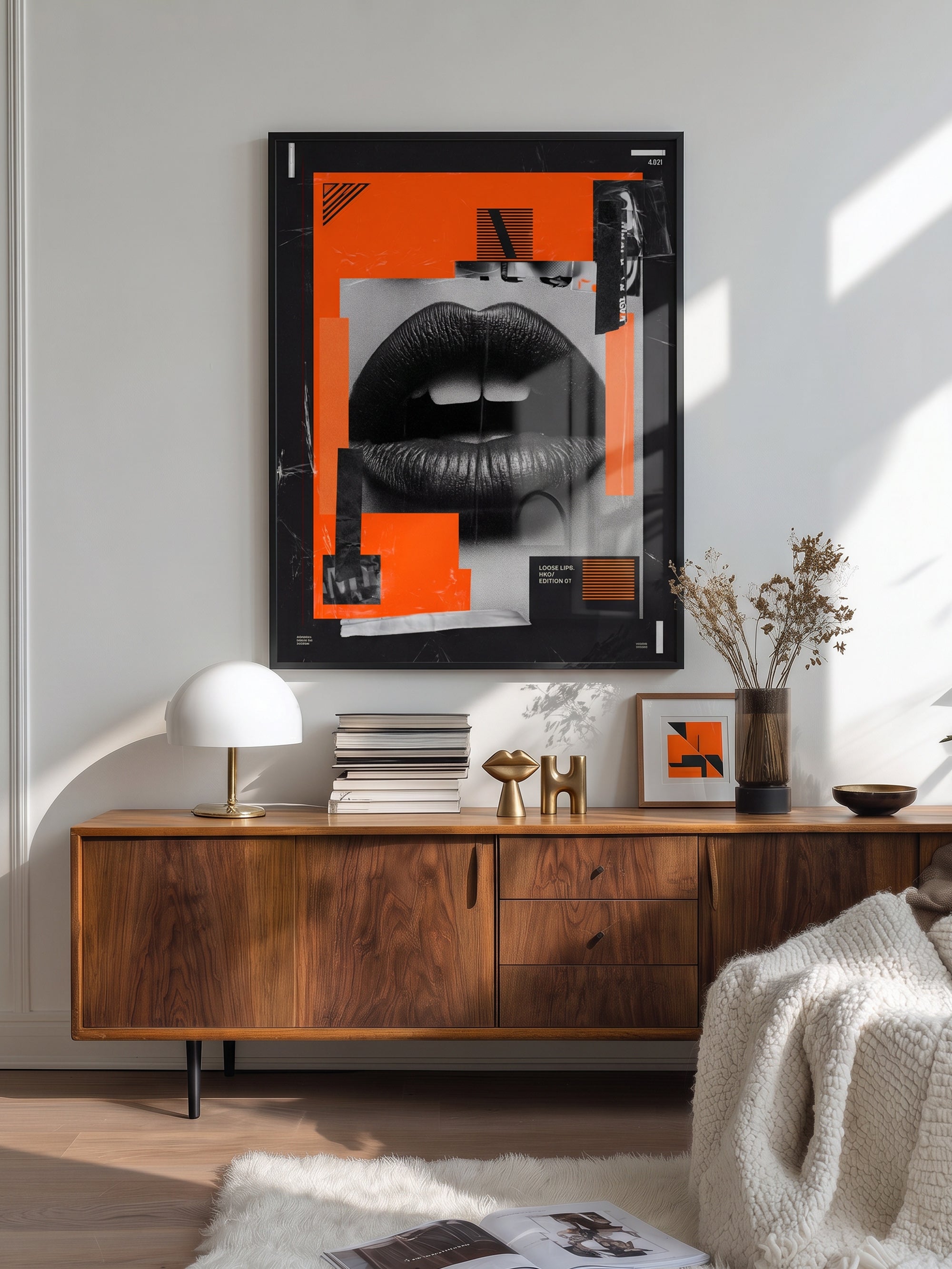 A framed abstract art print featuring black lips and bright orange geometric shapes, displayed on a light gray wall above a mid-century modern wooden sideboard in a styled living room.