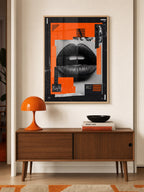 A framed abstract art print featuring black lips and bright orange geometric shapes, displayed on a beige wall above a mid-century modern wooden console table with an orange lamp and books.
